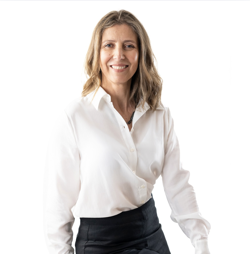 Woman seated on a stool against a plain light background, smiling at the camera, wearing a white blouse and dark trousers with one leg crossed.